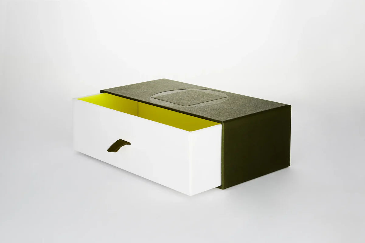 Three quarters profile view of Akoni Eyewear case storage box, featuring white cardboard drawer with a yellow interior and green fabric pull tab detail that slots into a green enclosure matching the leather case.