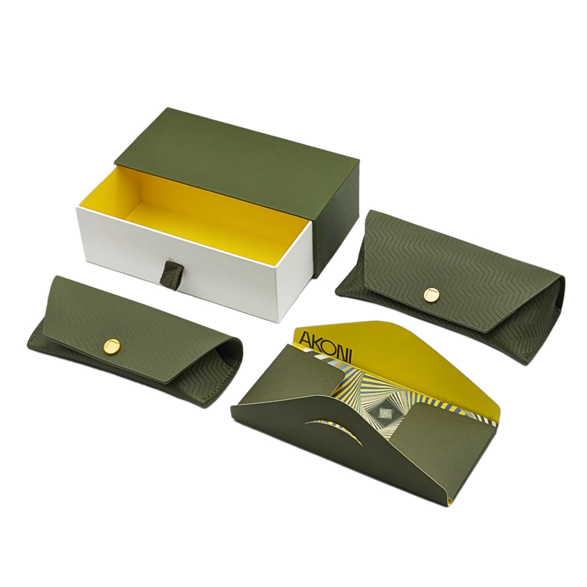 Three quarters profile view of Akoni case acessories, featuring one green case storage box with pull out drawer with white exterior, yellow interior, and green fabric pull tab detail, one large Akoni leather case, one small Akoni leather case, and one microfiber cleaning cloth in a paper envelope enclosure.