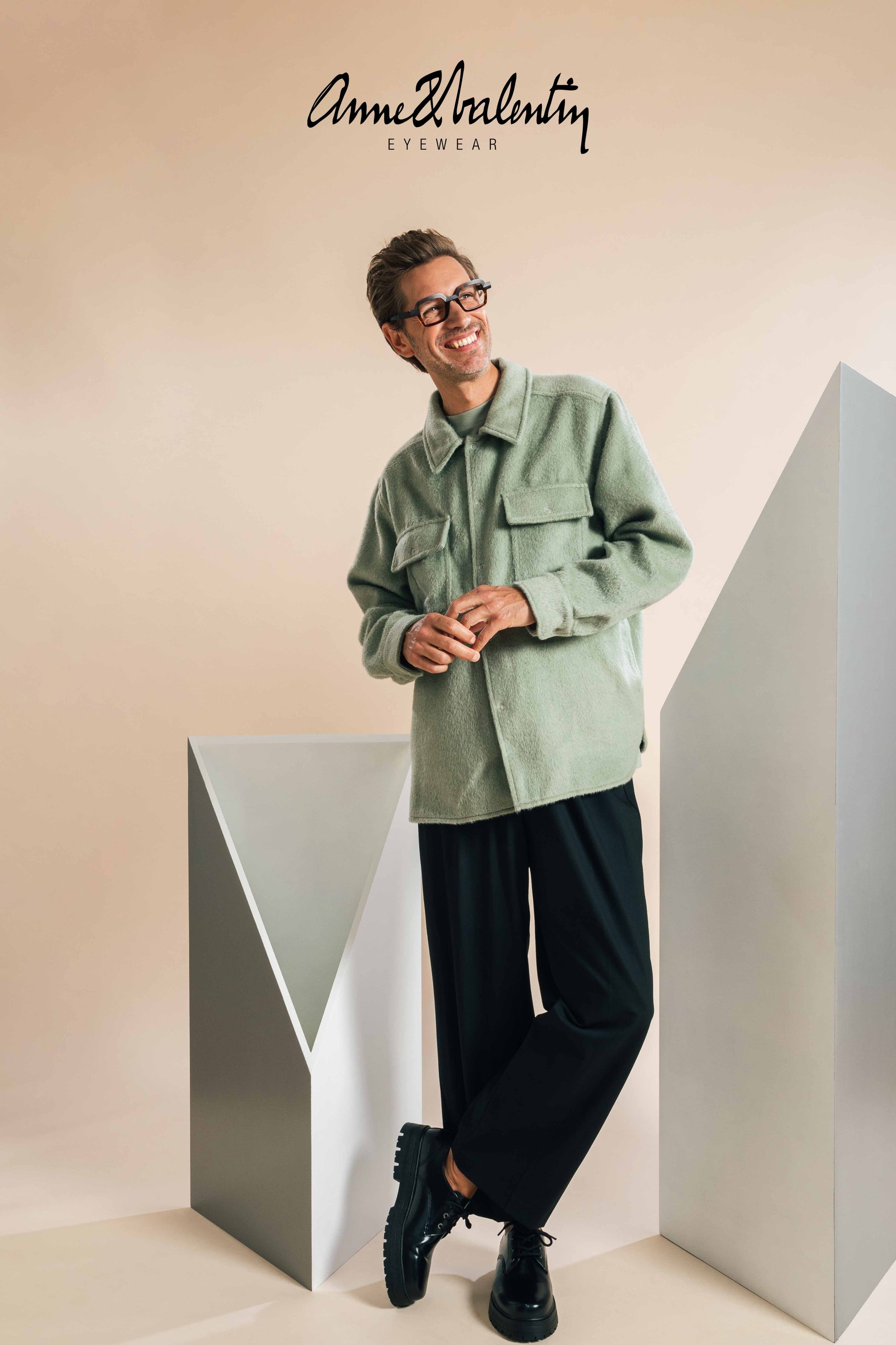 Person wearing a light green jacket and black pants standing between two geometrically shaped objects with 'Anne & Valentin' branding.