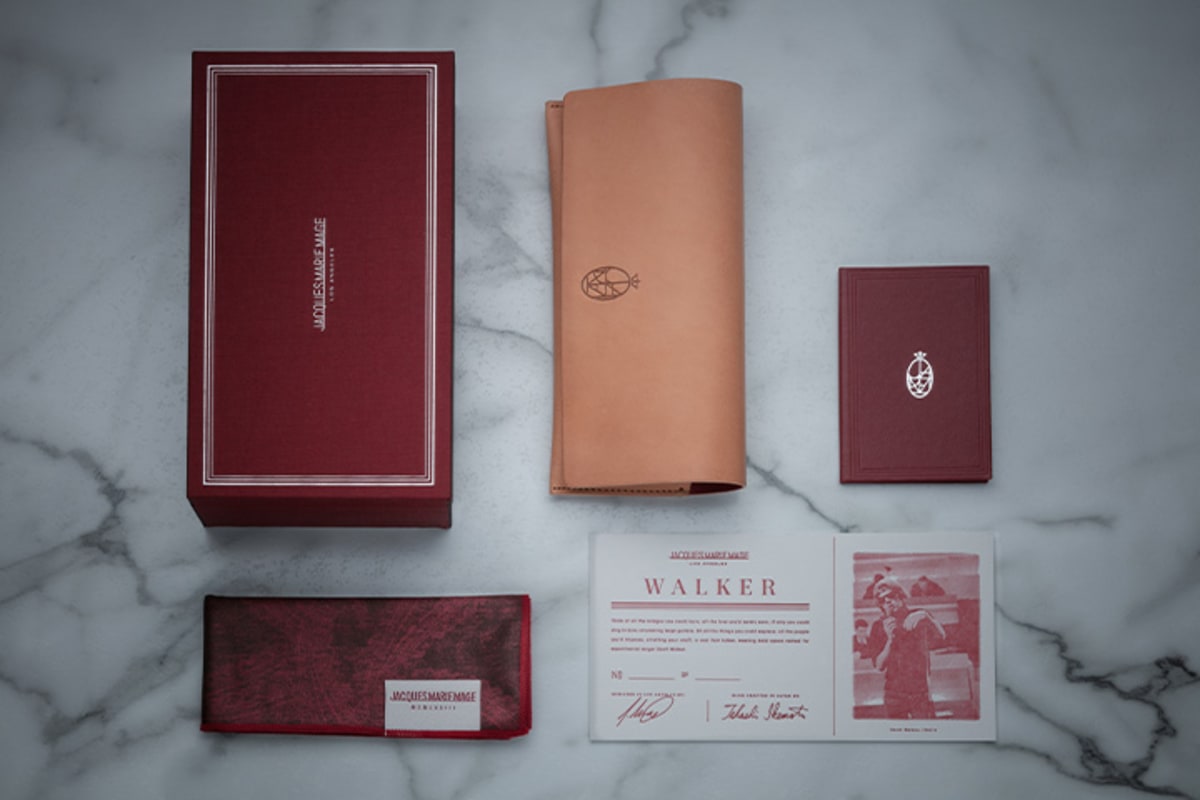 Jacques Marie Mage signature burgundy packaging and accessories, featuring Italian-handcrafted leather case, microfiber cleaning cloth/pocket square, silk fabric-covered packaging, and letterhead press certificate with limited edition serial number and craftsmen signature.